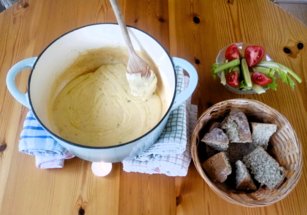 Cheese fondue in a blue enamel pan, stirred by a wooden spoon, supported over lit tealights by two stacks of tea-towels, next to a basket of wholemeal bread and a glass bowl of tomato, cucumber and celery salad on a wooden table